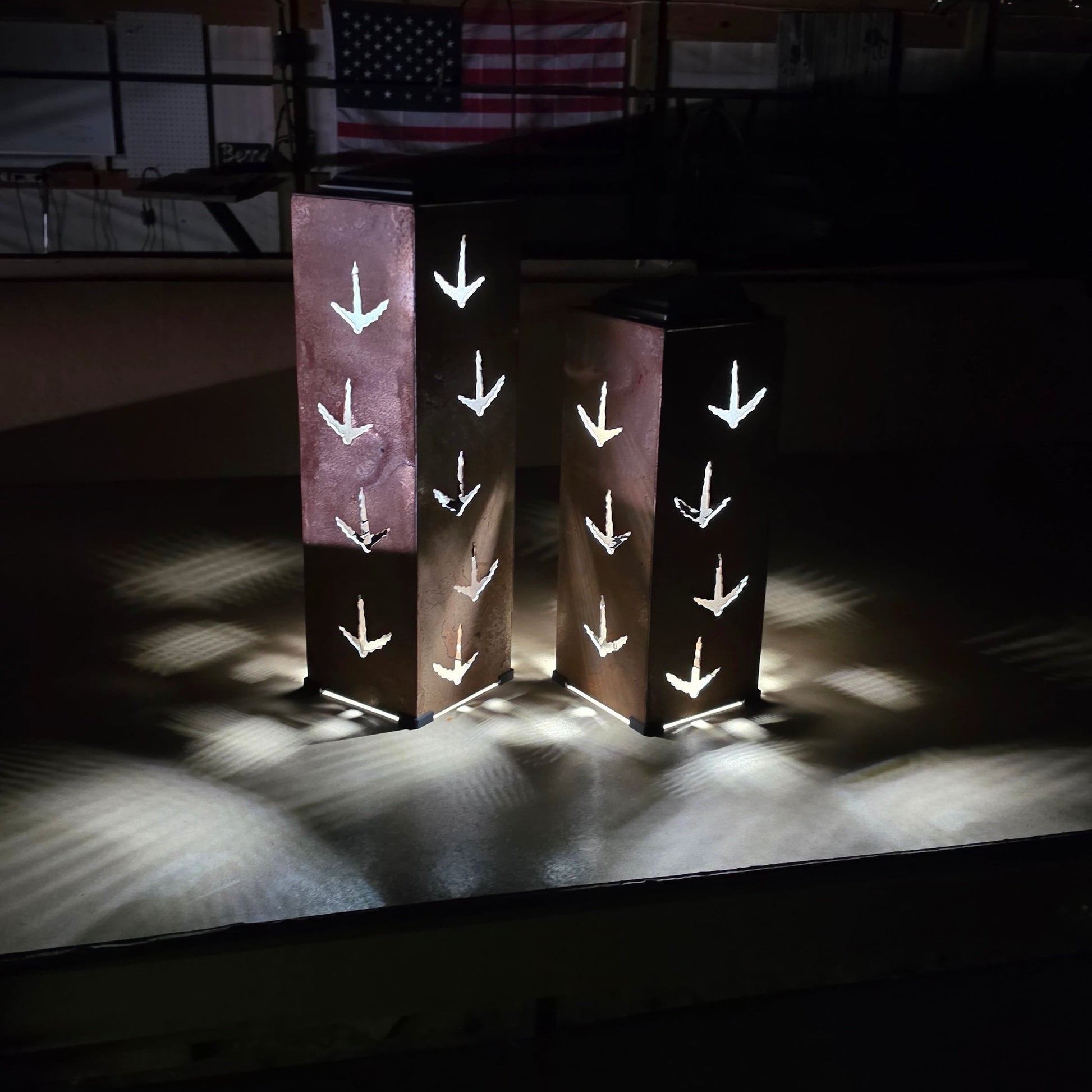 Two tall rectangular metal lanterns with cutout turkey print patterns, glowing with warm light on a dark stage-like setting with an American flag in the background.
