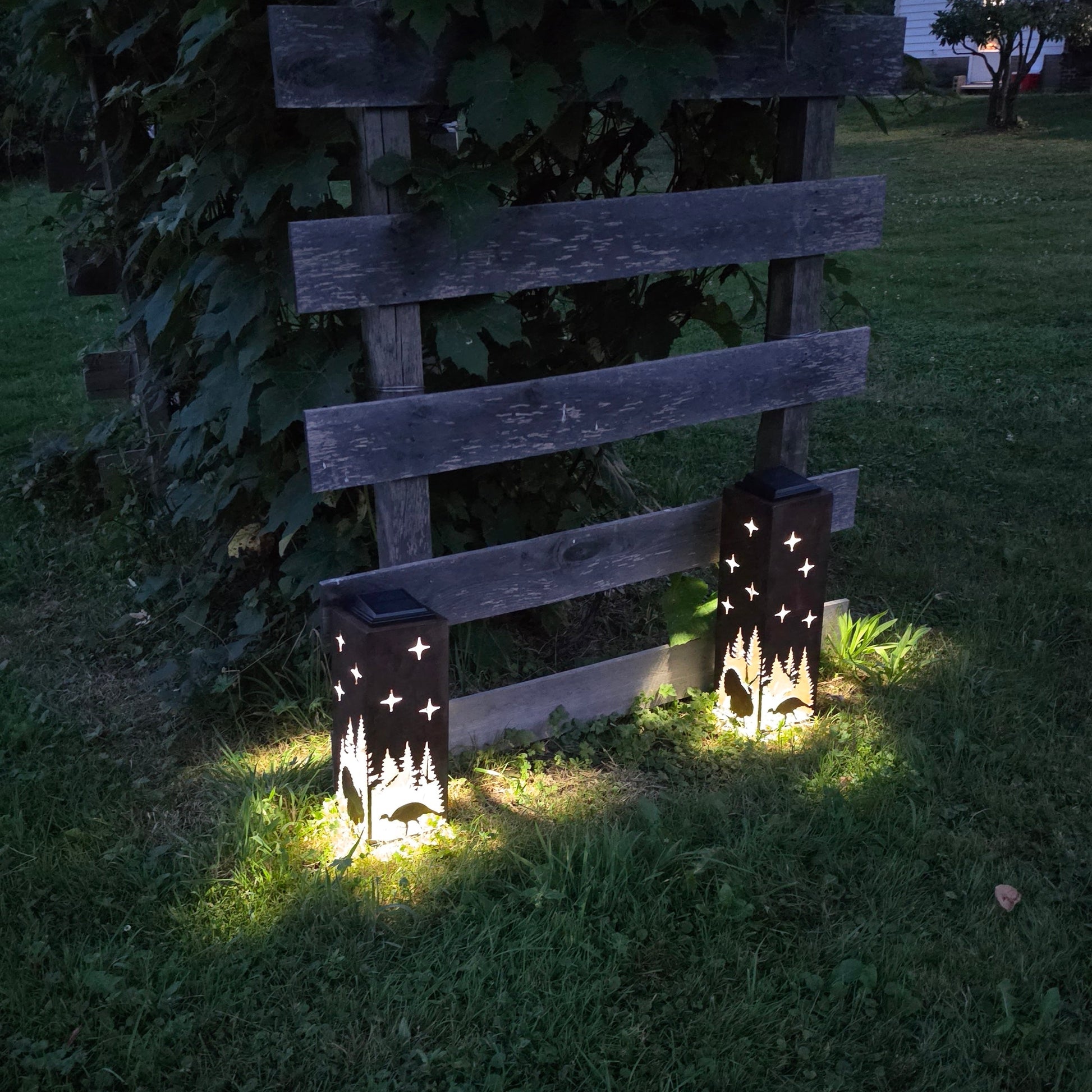 Two rustic solar pillars glowing at dusk in front of a grape vine with turkey designs