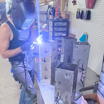A person wearing a welding helmet and protective gloves welds metal rectangular panels on a workbench in a workshop. The metal pieces have cutout star and crescent moon shapes.