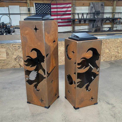 Two tall rectangular metal lanterns with a rustic copper-brown finish, featuring black cutout silhouettes of witches flying on broomsticks under a crescent moon and stars. The lanterns are photographed in a workshop setting with a concrete floor and wooden ceiling beams.