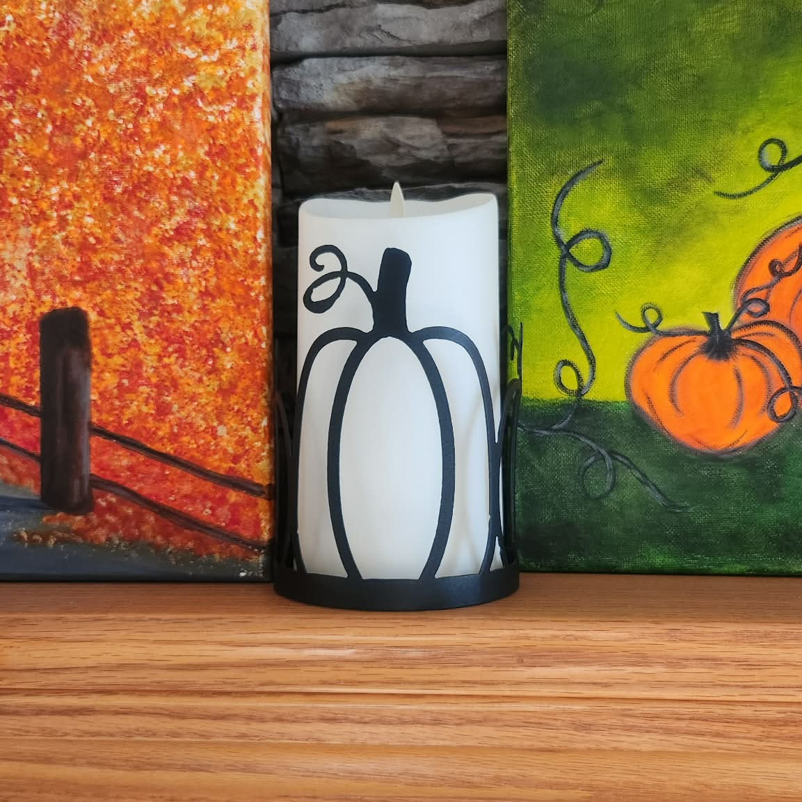 Candle with pumpkin design in front of autumn-themed paintings on a stone wall.